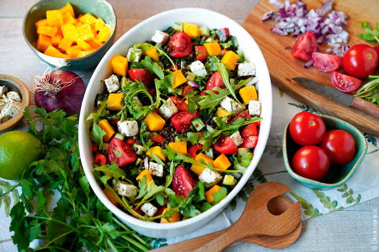 KI generiert: Ein bunter Salat mit Tomaten, Mango, Feta und Rucola in einer Schüssel, umgeben von frischen Zutaten.