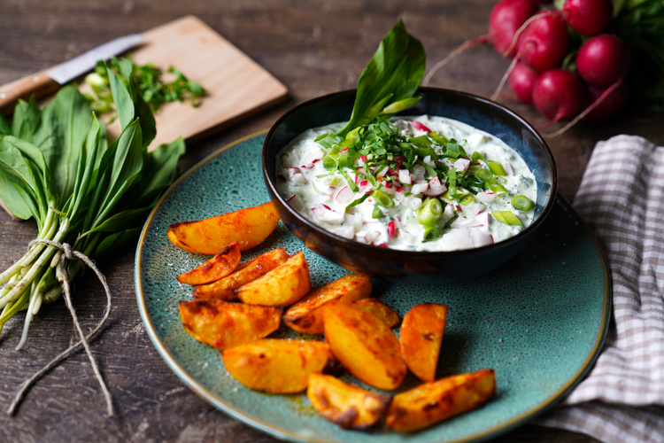 KI generiert: Gebratene Kartoffeln mit Kräuterdip, garniert mit Radieschen. Frische Zutaten wie Bärlauch liegen daneben.