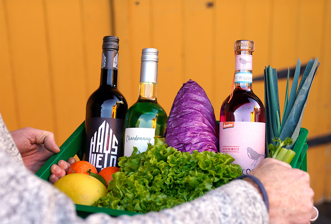 KI generiert: Ein Korb voller frischer Lebensmittel, darunter Rotkohl, Lauch, verschiedene Früchte und drei Flaschen Wein, wird von einer Person getragen. Der Hintergrund ist einfarbig gelb.