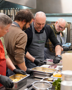 KI generiert: Das Bild zeigt mehrere Personen, die in einer Küche zusammenarbeiten, vermutlich bei einem Kochkurs oder einer Gruppenkochaktivität. Sie tragen Schürzen und sind mit der Zubereitung von Zutaten beschäftigt.