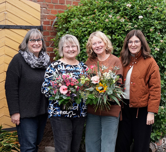 KI generiert: Vier Personen posieren lächelnd mit Blumensträußen vor einer grünen Hecke.