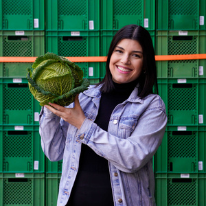 KI generiert: Eine Frau hält einen Wirsingkohl lächelnd vor einer Wand aus grünen Behältern. Der Hauptinhalt des Bildes ist die Präsentation des Kohls in einem fröhlichen Kontext.