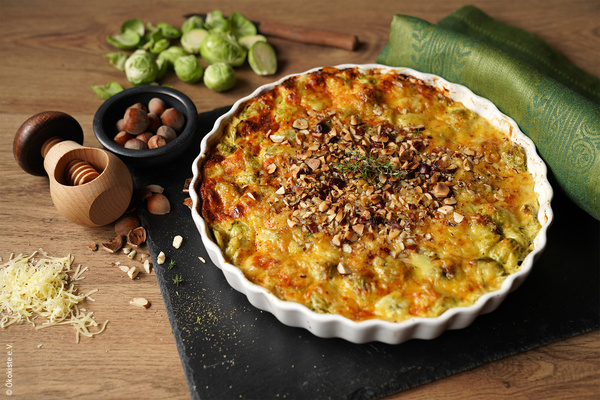 KI generiert: Ein Auflauf mit Nüssen, umgeben von Rosenkohl, Nüssen, Gewürzmühle und Käse. (© Ökokiste e.V.)