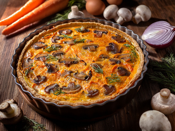 KI generiert: Eine Gemüsequiche mit Pilzen, Karotten und Kräutern auf einem Tisch mit weiteren Zutaten.