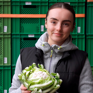 KI generiert: Eine Frau hält einen Wirsingkohl lächelnd vor einer Wand aus grünen Behältern. Der Hauptinhalt des Bildes ist die Präsentation des Kohls in einem fröhlichen Kontext.