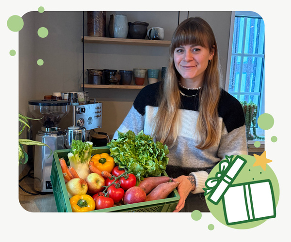 KI generiert: Zwei Personen stehen in einem Gewächshaus voller Pflanzen und halten eine Kiste mit frischem Gemüse. Der Fokus liegt auf der Präsentation der Ernte.