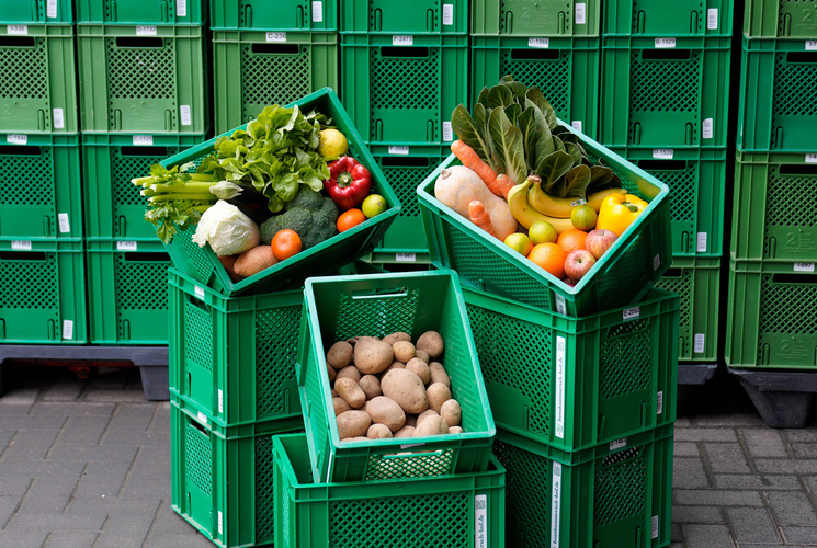 KI generiert: Grüne Kisten mit Gemüse und Früchten, darunter Kartoffeln, Paprika und Bananen, vor gestapelten Kisten.