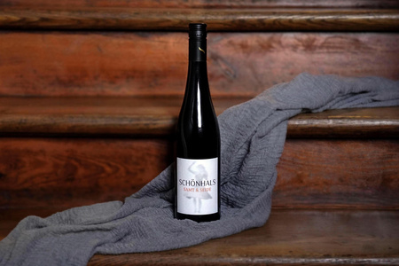 KI generiert: Das Bild zeigt eine Weinflasche mit der Aufschrift "Schönhals Samt & Seide" auf einer hölzernen Treppe, die von einem grauen Stoff umgeben ist. Der Fokus liegt auf der Weinflasche.