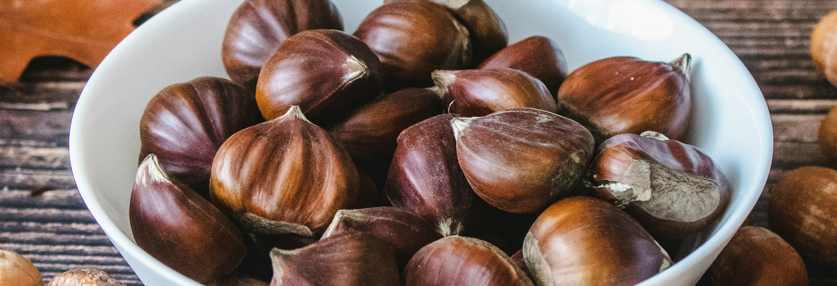 KI generiert: Das Bild zeigt eine Schüssel voller Kastanien, die auf einem Holztisch steht. Die Hauptinhalte sind die glänzenden, braunen Kastanien.