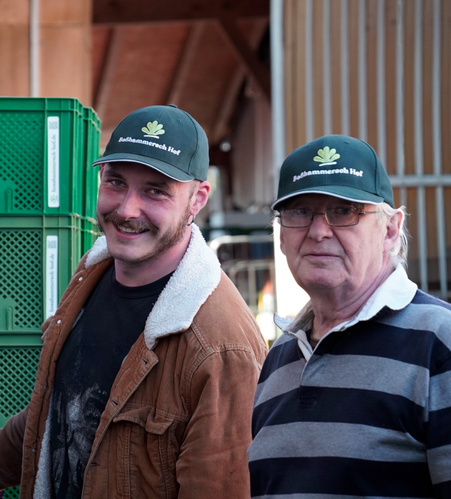 KI generiert: Zwei Männer mit grünen Kappen posieren vor grünen Kisten. Text auf Kappen: "Baßhammerc Hof".