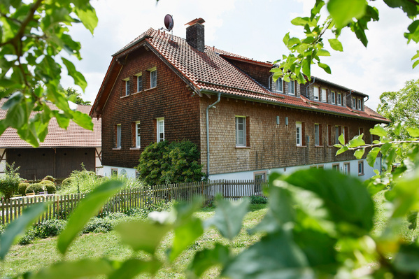 KI generiert: Holzbauernhaus in ländlicher Umgebung mit Garten und Zaun, umgeben von grüner Vegetation.