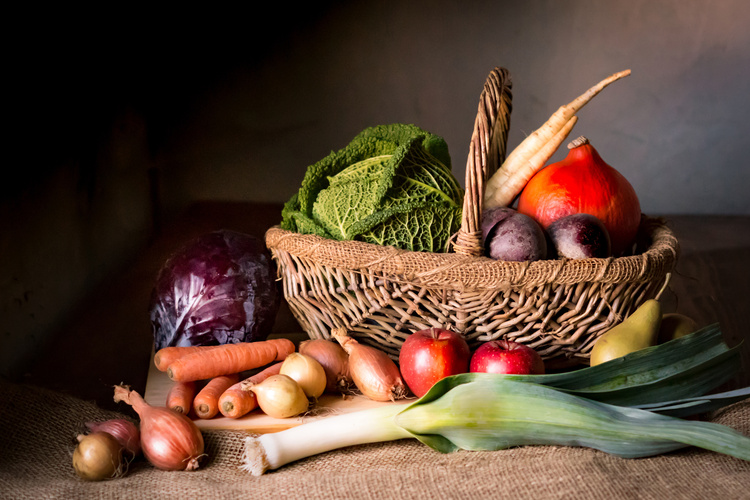 KI generiert: Ein Korb voller Gemüse und Obst wie Kohl, Karotten, Lauch und Äpfel auf rustikalem Hintergrund.