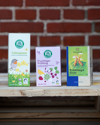 KI generiert: Drei Teepackungen vor einer Ziegelwand: "Frühlingsbote", "Fruchtiger Frühling", "Frühlings Kuss".