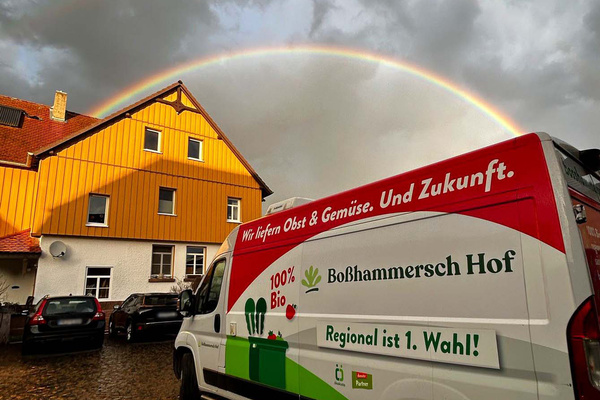 KI generiert: Lieferwagen vor gelbem Haus mit Regenbogen. Text: "Wir liefern Obst & Gemüse. Und Zukunft. 100% Bio. Regional ist 1. Wahl!"