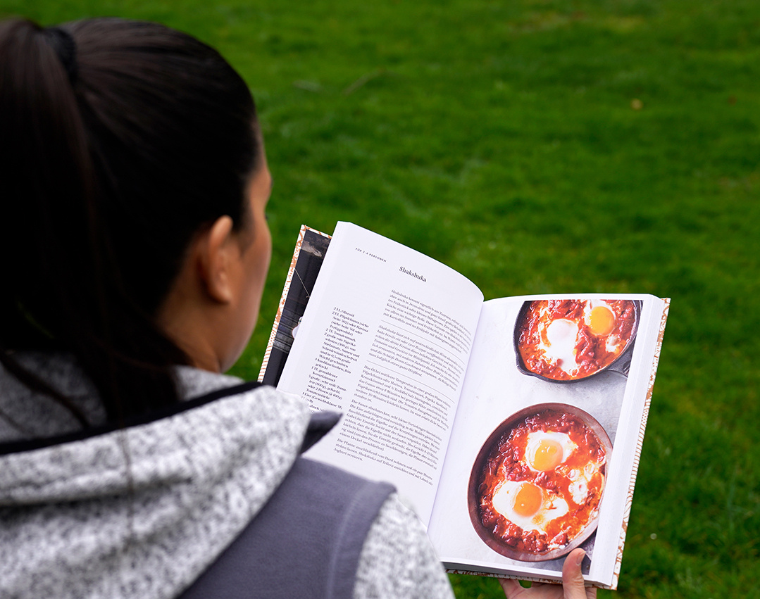 KI generiert: Eine Person hält ein Kochbuch in der Hand und betrachtet ein Rezept für Shakshuka, während sie im Freien steht. Das Hauptaugenmerk des Bildes liegt auf dem geöffneten Buch und den dargestellten Speisen.