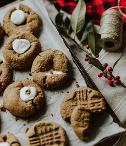 KI generiert: Das Bild zeigt eine Platte mit verschiedenen selbstgebackenen Keksen, die auf Backpapier liegen. Daneben liegen Dekorationselemente wie rote Beeren und grüne Blätter.