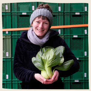 KI generiert: Eine Person hält ein Bündel frischen Pak Choi vor grünen Kisten. Sie lächelt in die Kamera.
