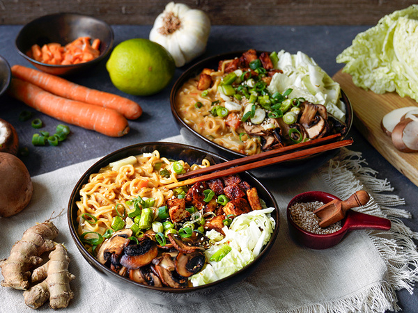 KI generiert: Zwei Schüsseln Ramen mit Gemüse und Tofu, umgeben von frischen Zutaten wie Karotten, Limette und Knoblauch.