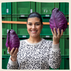 KI generiert: Eine lächelnde Frau hält zwei große Köpfe roten Spitzkohl in den Händen. Im Hintergrund sind gestapelte grüne Kisten zu sehen.
