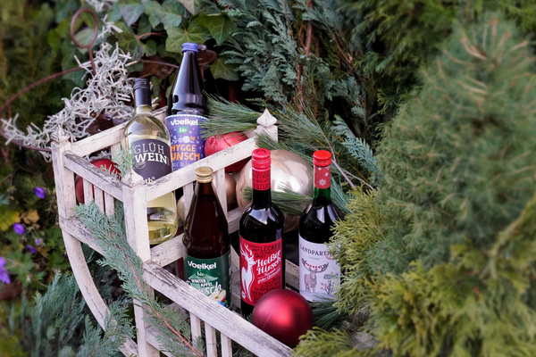 KI generiert: Getränke in einem Holzkorb mit Weihnachtsdekoration. Text: „Glühwein“, „Voelkel“, „Bioglühpunsch“.