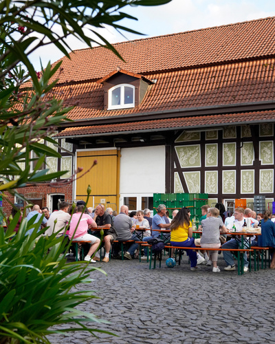 KI generiert: Menschen sitzen an Tischen im Freien vor einem Fachwerkhaus und unterhalten sich.