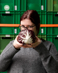 KI generiert: Eine Frau hält einen Wirsingkohl lächelnd vor einer Wand aus grünen Behältern. Der Hauptinhalt des Bildes ist die Präsentation des Kohls in einem fröhlichen Kontext.