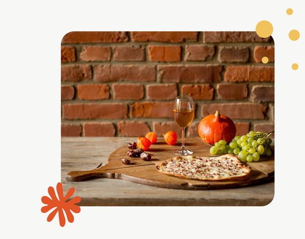 KI generiert: Ein Flammkuchen auf Holzbrett mit Wein, Kürbis, Trauben und Kastanien vor einer Backsteinwand.