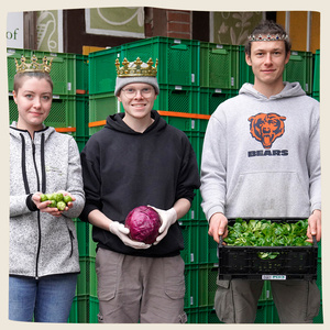 KI generiert: Drei Personen tragen Kronen und halten Gemüse vor einem Hintergrund aus grünen Kisten. Jede Person präsentiert stolz ein anderes Gemüse: Gurken, Rotkohl und Salat.