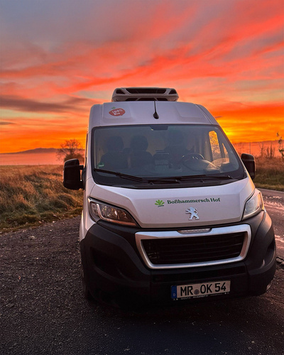 KI generiert: Ein Lieferwagen mit "Boßhammersch Hof"-Logo vor einem dramatischen Sonnenuntergang.