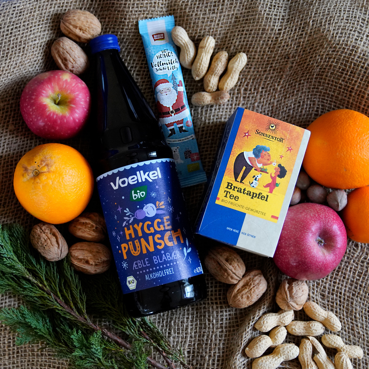 KI generiert: Das Bild zeigt eine Auswahl von Herbst- und Winterprodukten, darunter eine Flasche Hygge Punsch, Bratapfeltee, Orangen, Äpfel, Nüsse und eine Schokoladenfigur. Die Gegenstände sind auf einem groben Stoff drapiert.