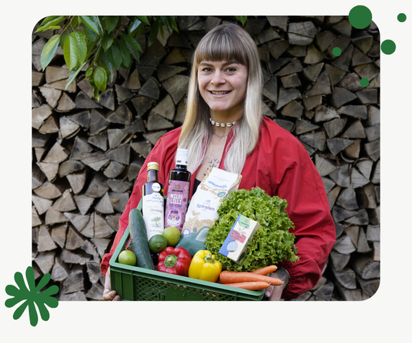 KI generiert: Zwei Frauen stehen vor einer Holzwand; eine von ihnen hält eine grüne Kiste mit verschiedenen Lebensmitteln wie Gemüse und Flaschen. Das Bild vermittelt einen ländlichen oder marktorientierten Kontext.