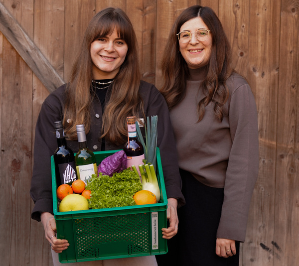 KI generiert: Zwei Frauen stehen vor einer Holzwand; eine von ihnen hält eine grüne Kiste mit verschiedenen Lebensmitteln wie Gemüse und Flaschen. Das Bild vermittelt einen ländlichen oder marktorientierten Kontext.