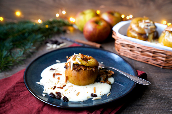 KI generiert: Das Bild zeigt einen gebackenen Apfel auf einem blauen Teller, serviert mit Vanillesoße und dekoriert mit Nüssen und Rosinen. Im Hintergrund sind weitere Äpfel in einem Korb sowie weihnachtliche Dekorationen zu sehen.