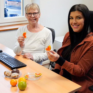 KI generiert: Zwei Personen sitzen an einem Tisch und lächeln, während sie Fruchtstücke halten. Auf dem Tisch liegen Gebäck und verschiedene Zitrusfrüchte.