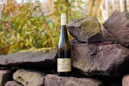 KI generiert: Das Bild zeigt eine Weinflasche mit der Aufschrift "Schönhals Samt & Seide" auf einer hölzernen Treppe, die von einem grauen Stoff umgeben ist. Der Fokus liegt auf der Weinflasche.