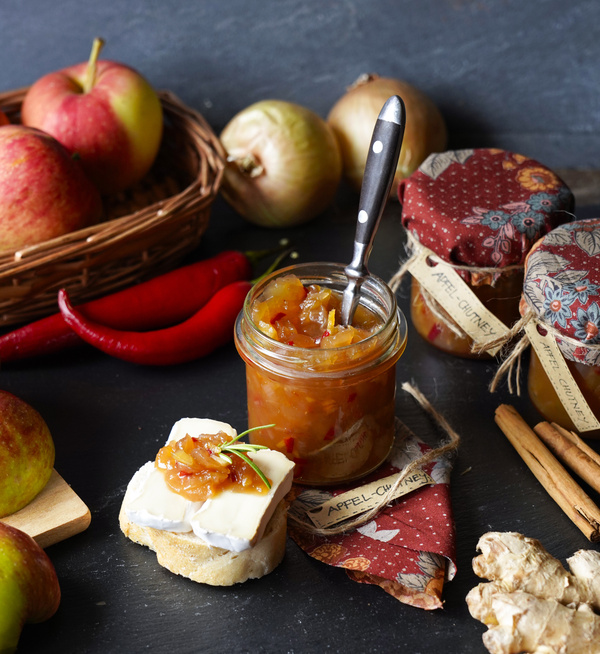 KI generiert: Ein Glas Apfel-Chutney umgeben von Äpfeln, Zwiebeln und Chilis; Text: "Apfel-Chutney".