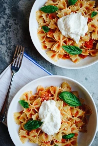 KI generiert: Das Bild zeigt zwei Schüsseln mit Farfalle-Nudeln, die mit Tomatensauce, frischem Basilikum und einem Klecks Ricotta garniert sind. Daneben liegt eine Gabel auf einer Serviette.
