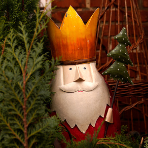 KI generiert: Das Bild zeigt eine weihnachtliche Figur mit Krone und Bart, die einen kleinen Tannenbaumstab hält. Die Figur ist von Pflanzen umgeben und hat einen festlichen, rustikalen Hintergrund.