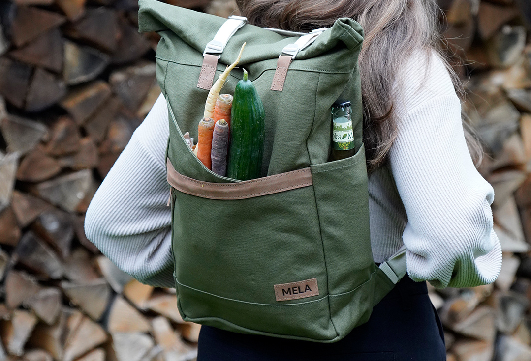 KI generiert: Eine Person trägt einen grünen Rucksack auf dem Rücken, in dem Gemüse und eine kleine Flasche zu sehen sind. Der Hintergrund besteht aus gestapeltem Feuerholz.