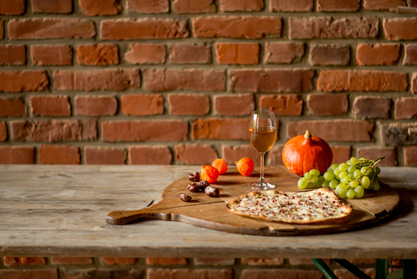 KI generiert: Herbstliches Stillleben mit Kürbis, Weintrauben, Flammkuchen und einem Glas Weißwein auf rustikalem Tisch.